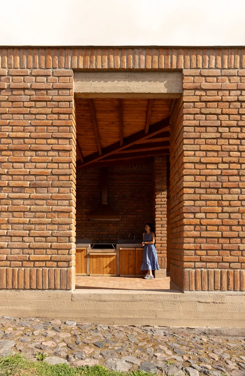 Wide brick portico with exposed timber ceiling sheltering a figure standing beside timber cabinetry