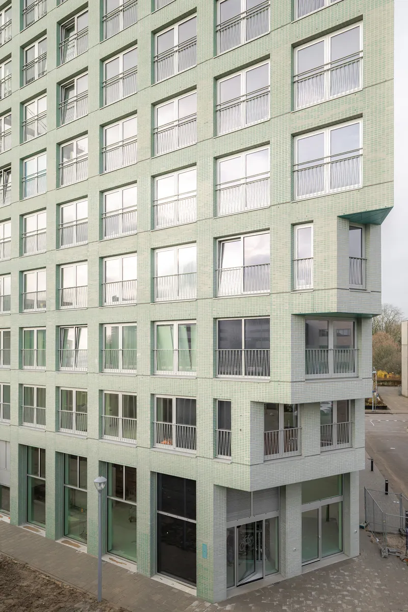 Corner view of the pale green tiled facade with balconies and ground-floor glazing on an overcast day