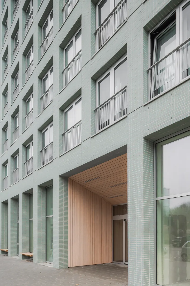 Street entrance with timber-lined soffit recessed into the pale green tiled facade below balconies with railings