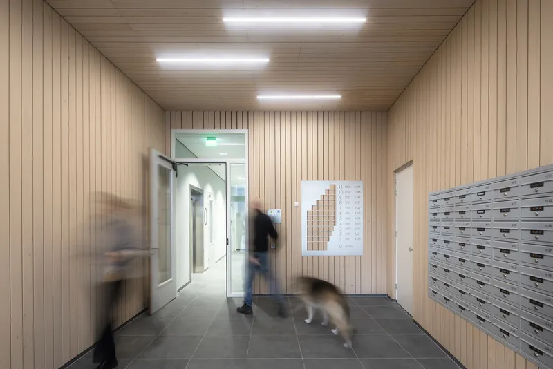 Timber-lined lobby with grey tile floor, mailboxes, and a person walking toward the glass door in motion blur