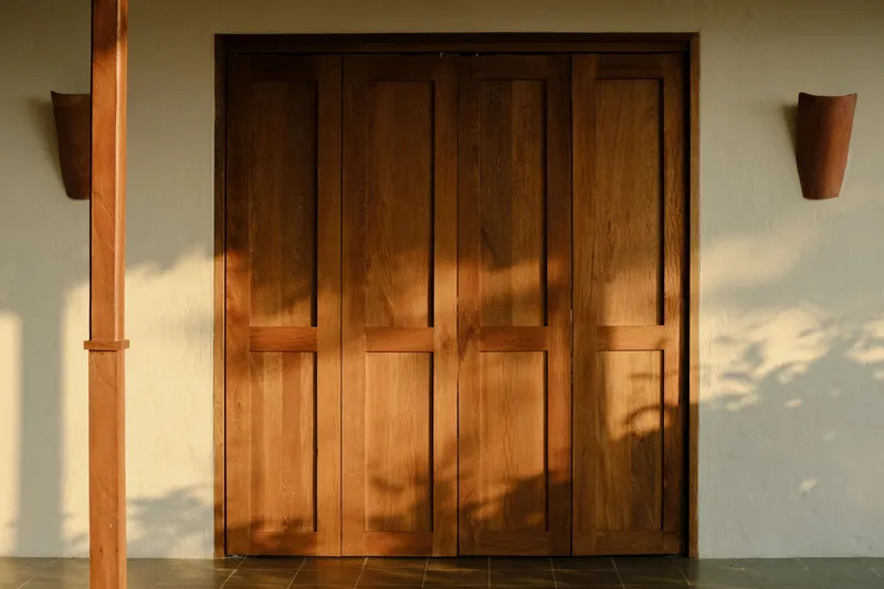 Four-panel timber folding doors beneath a wooden column with dappled tree shadows across the plastered wall