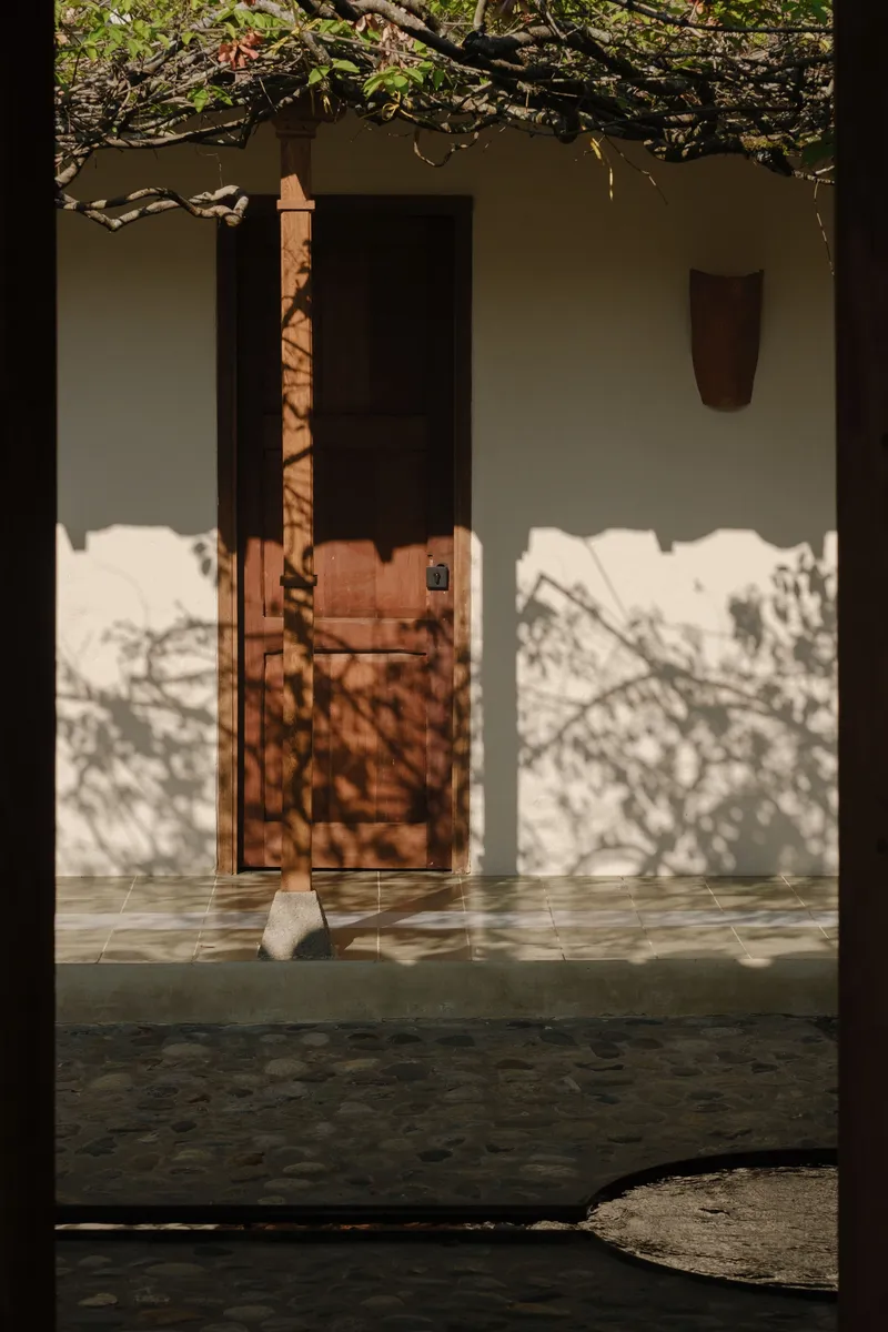 Covered porch with timber column and tiled floor seen through dark doorway with filtered tree shadows