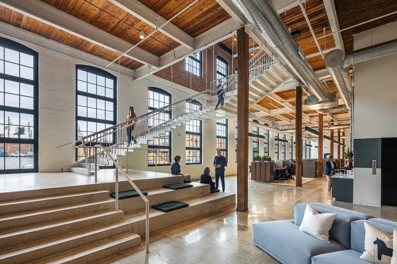 Multi-level lobby with steel and wood staircase, timber columns, black-framed windows and people gathering