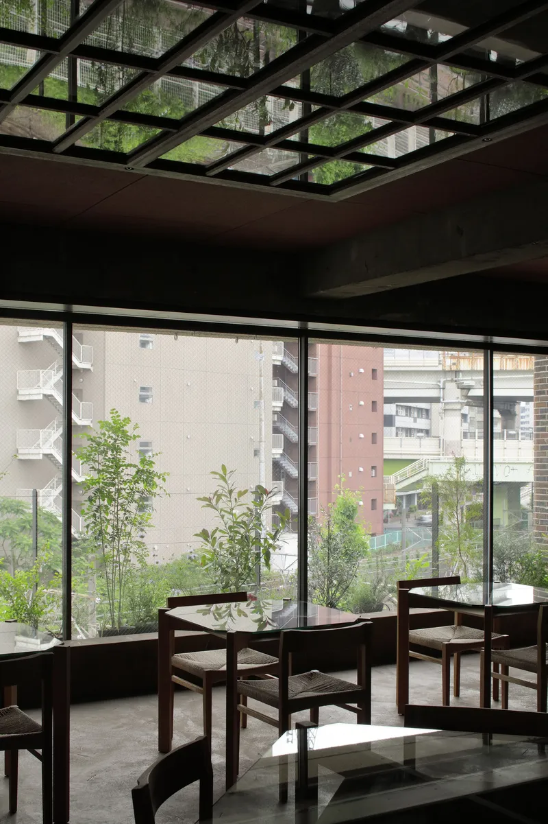 Dining area with black steel grid skylight overhead and floor-to-ceiling windows overlooking planted terrace and neighboring buildings