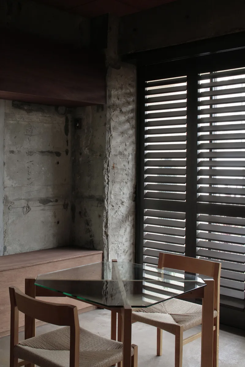 Interior corner showing exposed concrete walls with horizontal louvers filtering light beside timber and glass dining table
