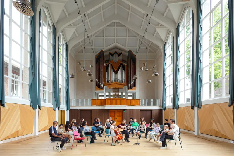 Tall vaulted hall with exposed timber ceiling structure, pipe organ balcony, and group seated in circle