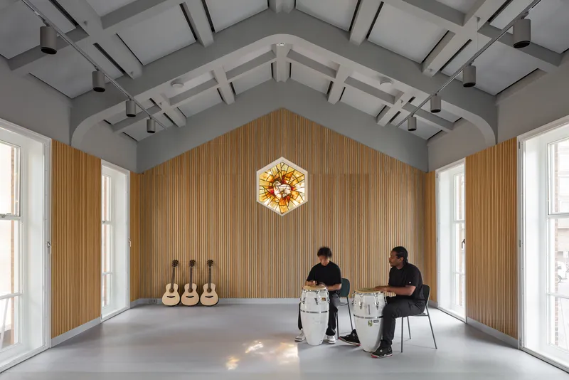 Vaulted rehearsal space with vertical timber wall cladding and two musicians playing hand drums