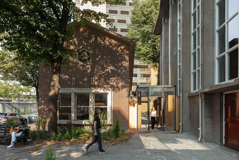 Brick gable end with ground-floor glazing viewed through dappled tree shade and planted beds