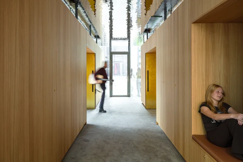 Corridor with light wood wall panels, yellow doorways, and people passing through under decorative ceiling installation