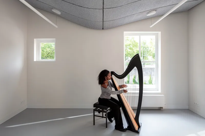 Practice room with curved acoustic ceiling panel, grey floor and musician playing harp by window