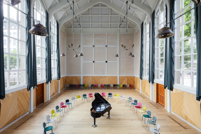Vaulted performance hall with grand piano surrounded by colorful seating under industrial pendant lights
