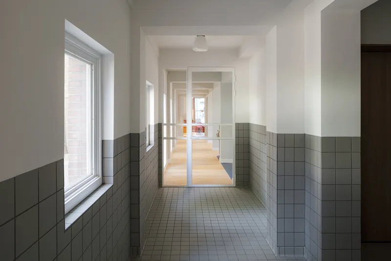 Corridor with grey tile wainscoting leading through glass doors toward daylit rooms beyond