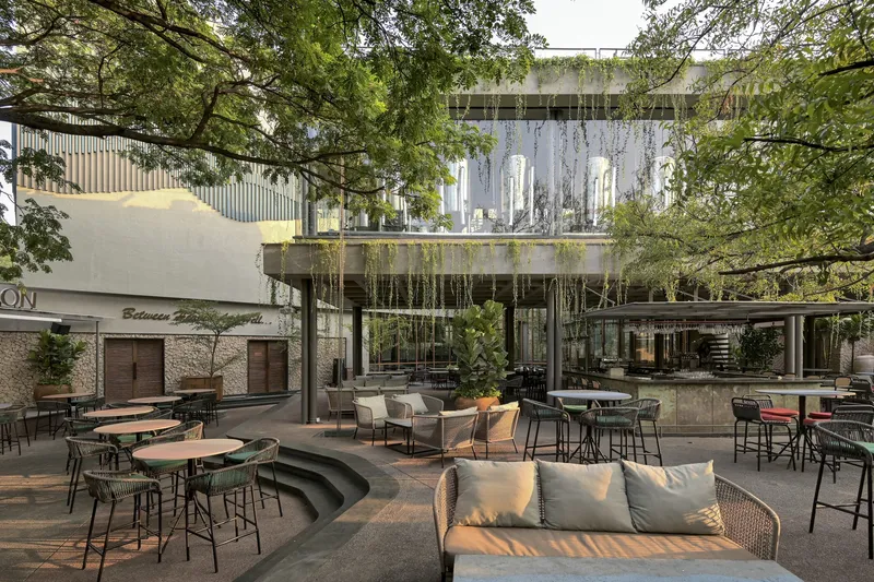 Outdoor courtyard with curved seating platform and cafe tables beneath overhanging planted terraces and surrounding trees
