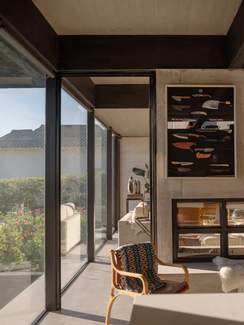 Interior corner with black steel framed glazing opening to a planted terrace in afternoon light