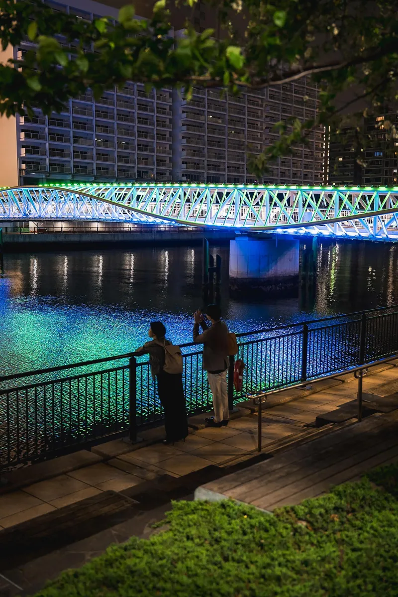 Two figures photographing the lit truss bridge from a waterfront promenade framed by foliage
