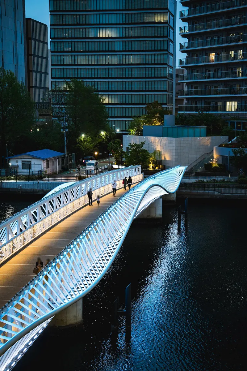 Illuminated pedestrian bridge with white lattice railings spanning a canal at dusk