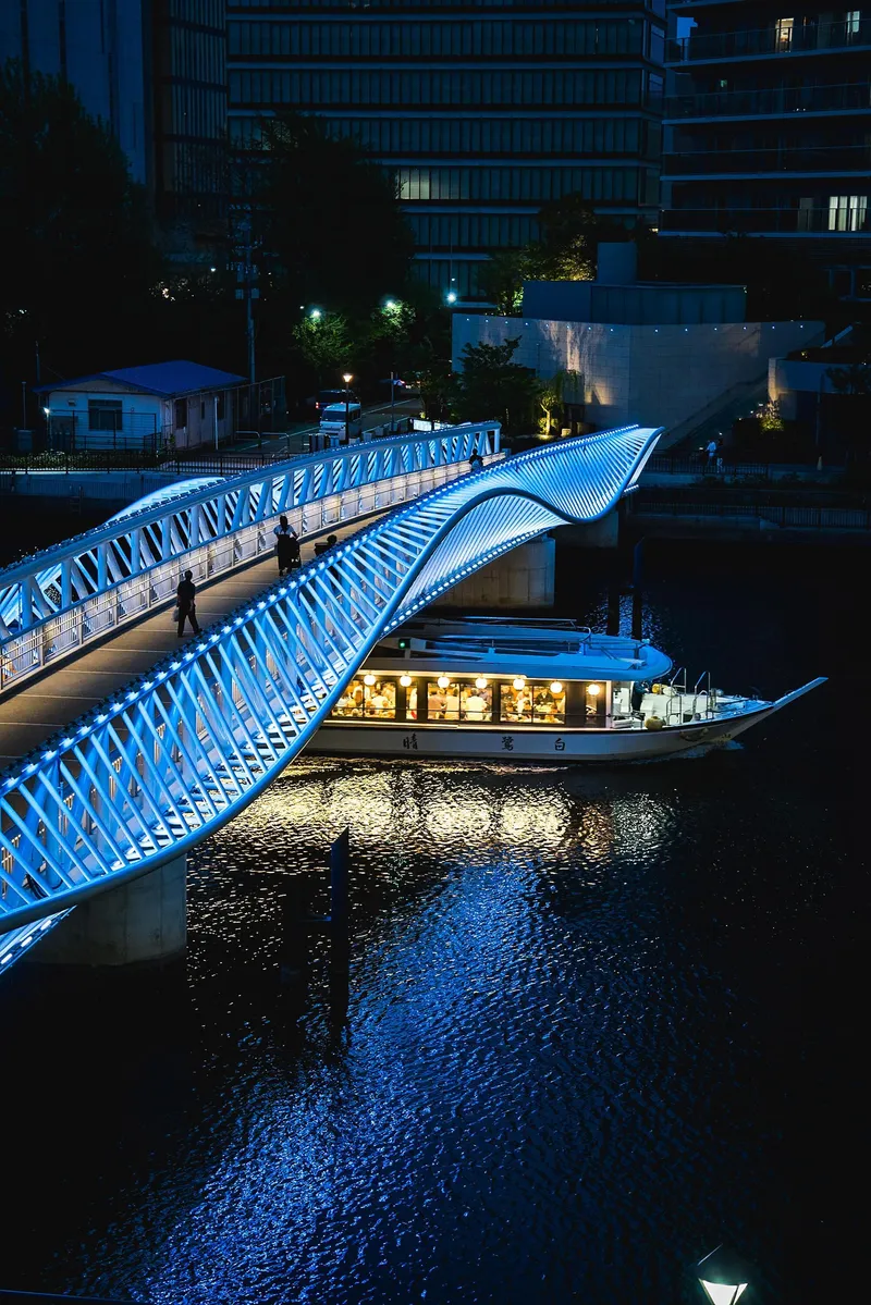 Illuminated curved bridge over waterway with a passing boat underneath at night