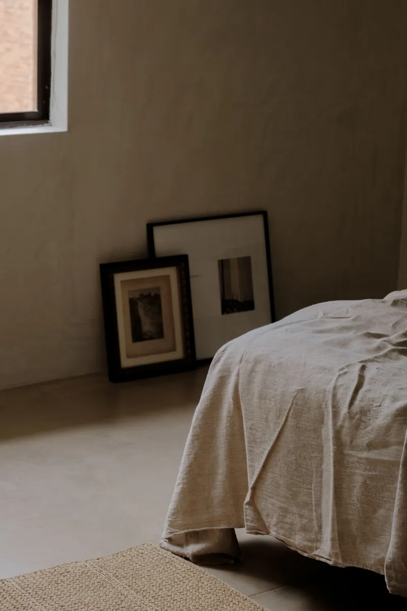 Bedroom corner with two framed artworks leaning against the plaster wall beside a linen-covered bed