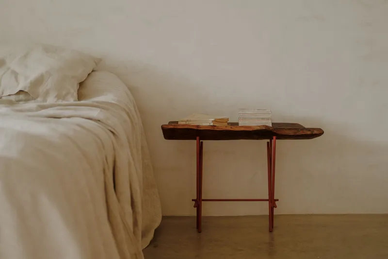 Live-edge timber side table with red metal legs beside a rumpled bed in warm daylight