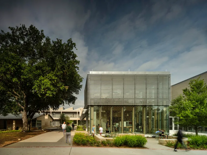 Glass box pavilion with horizontal sunshade louvers framed by a large tree and pedestrian pathway