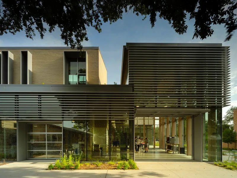Entrance facade with horizontal bronze louvers and planted bed beneath overhanging tree branches at dusk