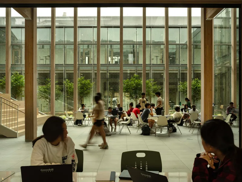 Double-height dining hall with tall timber-framed windows overlooking a planted courtyard and students studying at tables