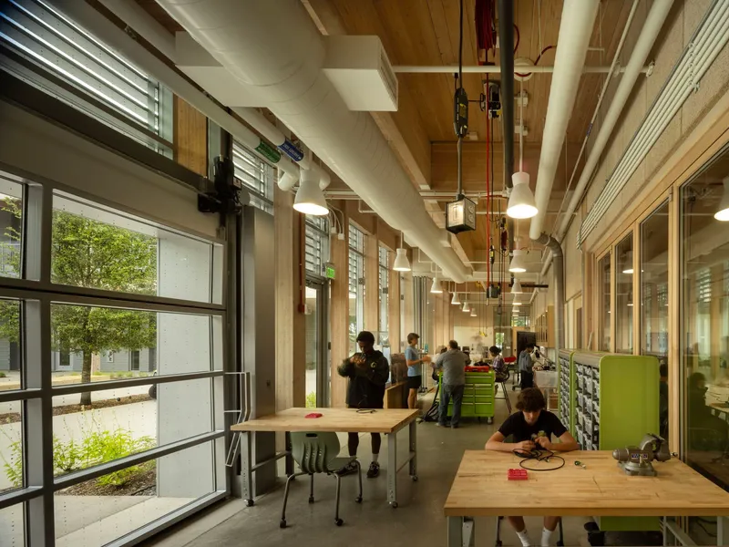 Workshop corridor with exposed timber ceiling, pendant lights, and workbenches along glazed walls with courtyard views
