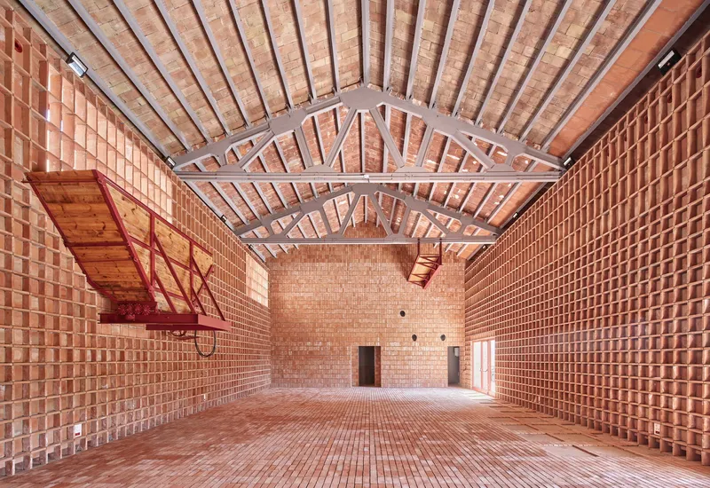 Interior hall with perforated terracotta brick walls, exposed timber trusses and a red steel balcony under diffused daylight
