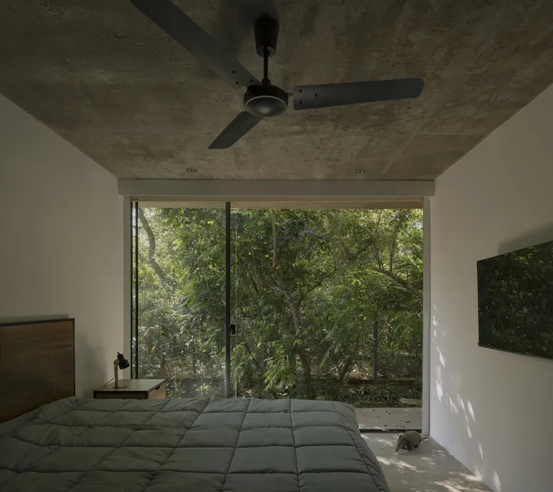 Bedroom with exposed concrete ceiling and ceiling fan facing full-height glazing opening to forested landscape