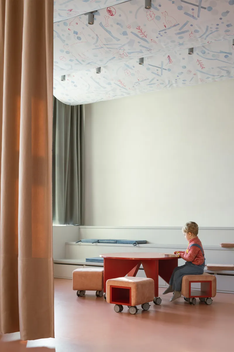 Child seated at red table beneath patterned fabric ceiling with beige and green curtains framing window