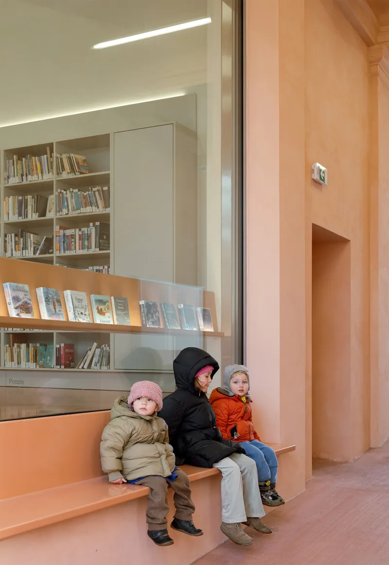 Three children in winter jackets seated on pink built-in bench beside glass display case