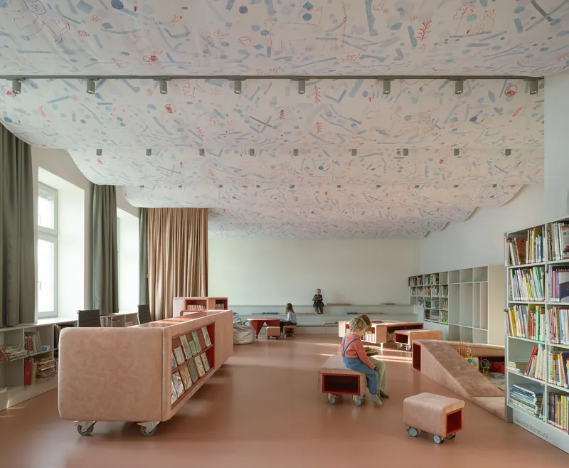 Library interior with patterned fabric ceiling, mobile pink shelving units, and children reading on a terracotta floor