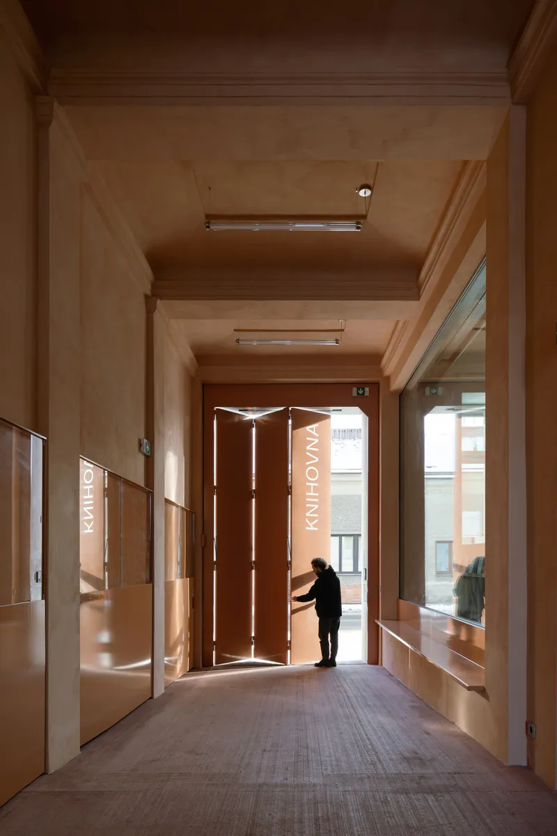 Entry vestibule with plywood walls and ceiling as a figure opens the folding bronze entrance door