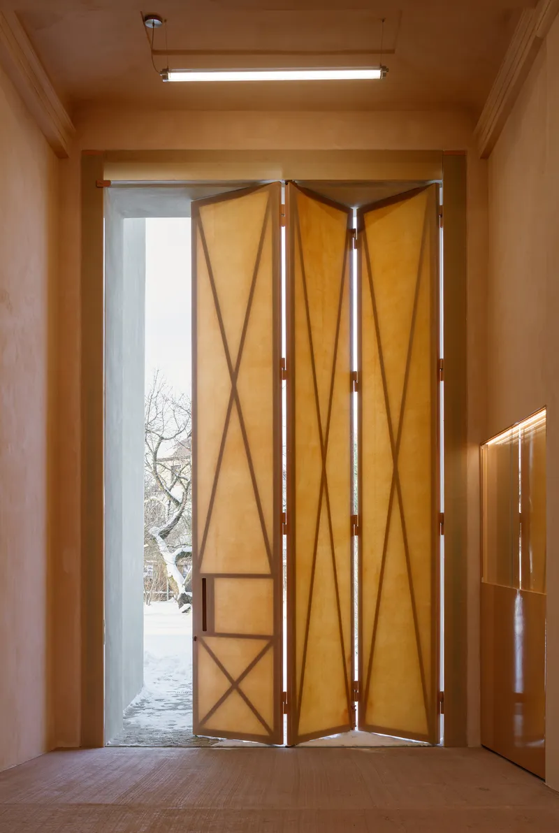 Folding timber door with diagonal bracing pattern opening to a snowy courtyard with bare tree