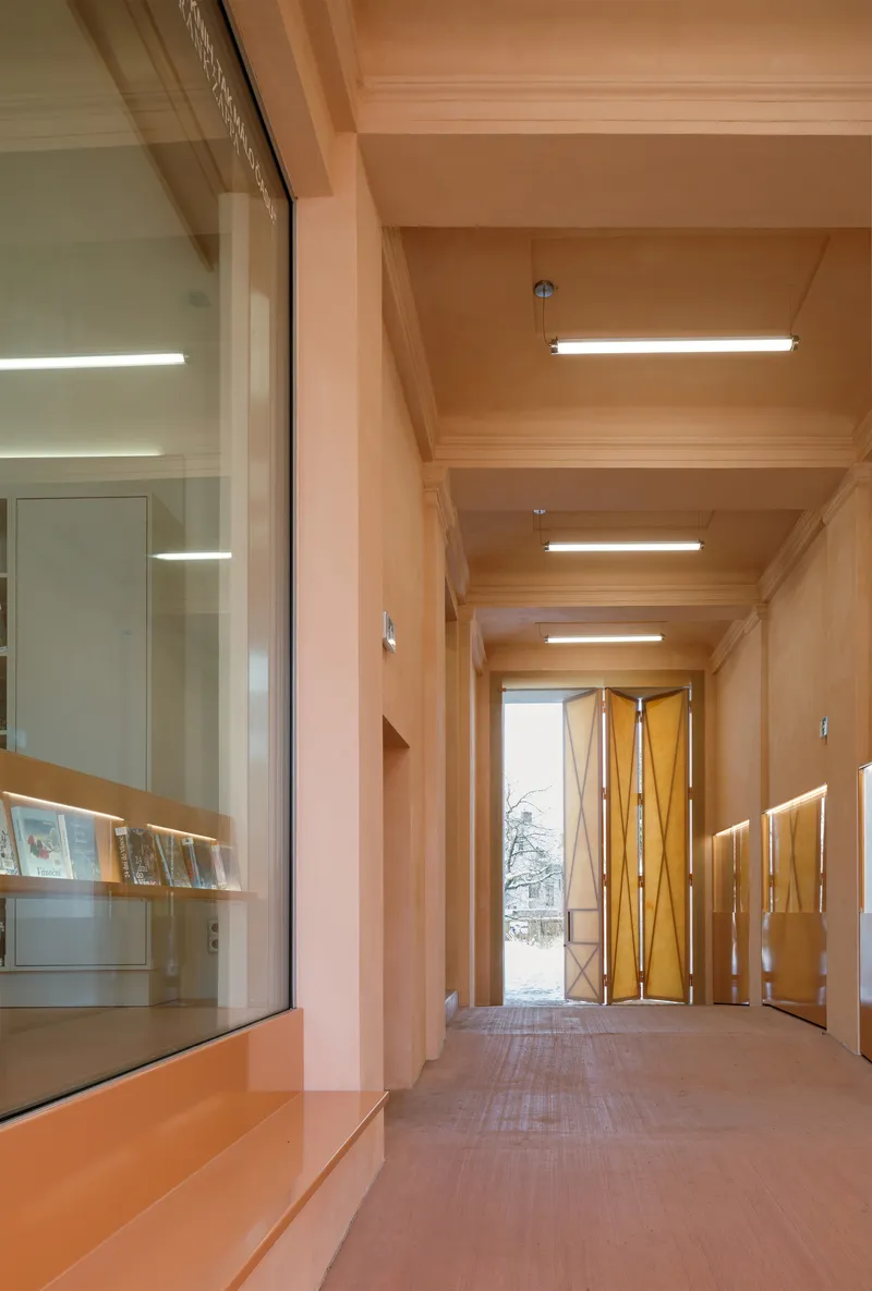 Interior corridor with exposed timber beams, linear ceiling lights, and folded copper entry doors at the end