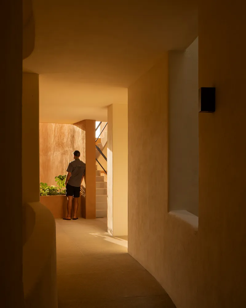 Interior passage with arched openings and warm light revealing a figure standing near a planted courtyard