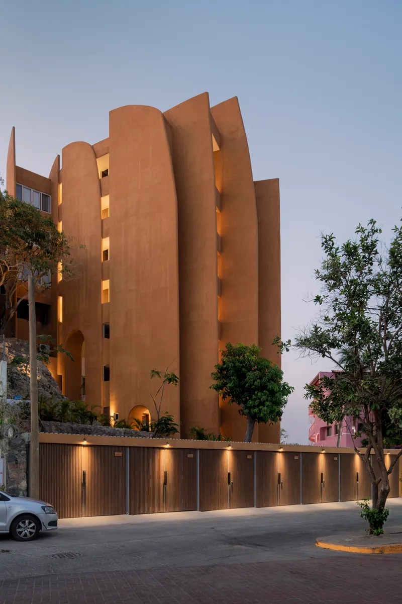 Curved ochre facade with illuminated balconies and a ribbed ground-level wall at twilight