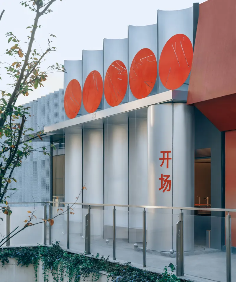 Curved white columns supporting red circular panels beside a glass railing and young tree