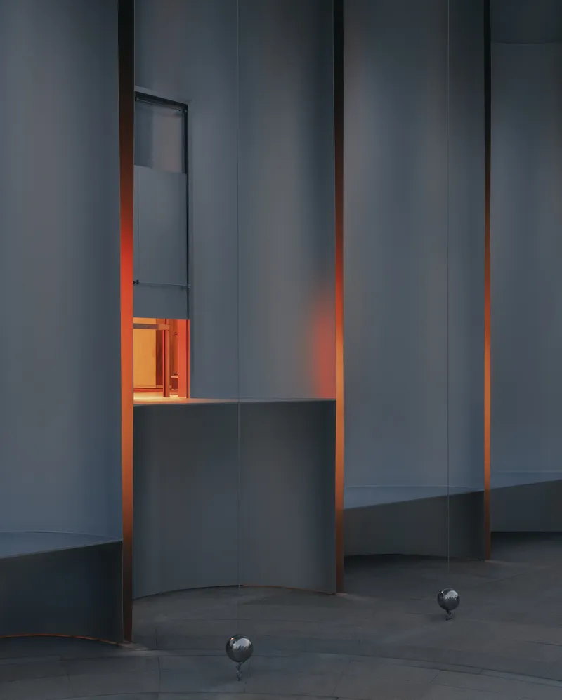 Vertical slot window framing warm interior light within grey paneled walls at dusk