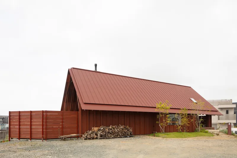 Red corrugated metal gable roof and walls surrounded by a gravel yard with stacked firewood