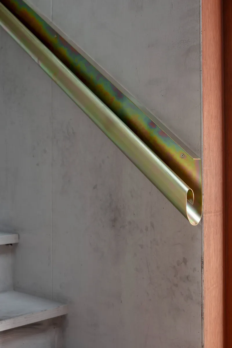 Detail of iridescent brass handrail mounted to grey concrete wall beside timber doorframe