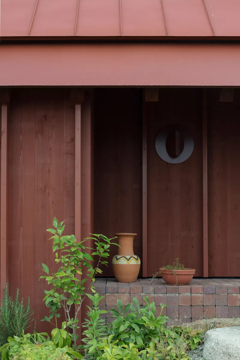 Timber slatted facade beneath a pink metal roof with potted plants on a tiled threshold