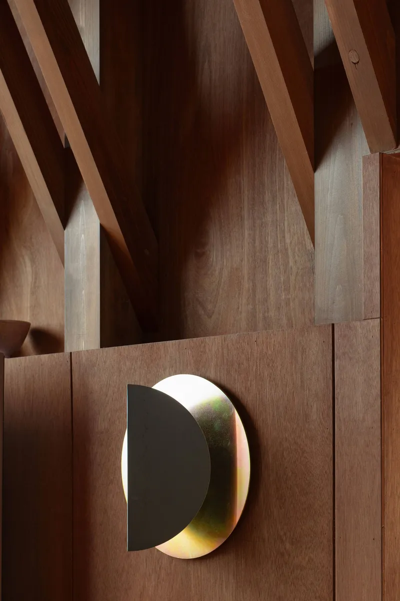 Circular brass wall sconce mounted on wood paneling beneath diagonal timber rafters in afternoon light