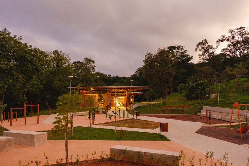 Community building with timber roof overlooking a plaza with young plantings and playground equipment at twilight