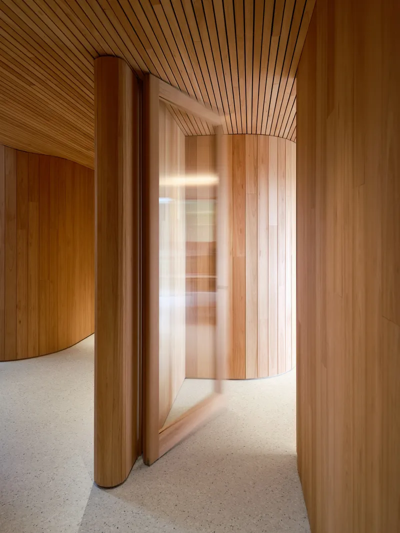 Curved timber corridor with slatted ceiling leading around cylindrical columns in natural daylight