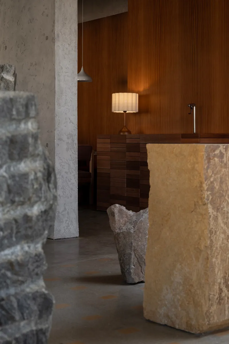 Vertical timber wall paneling behind a table lamp and rough stone column fragment on concrete floor