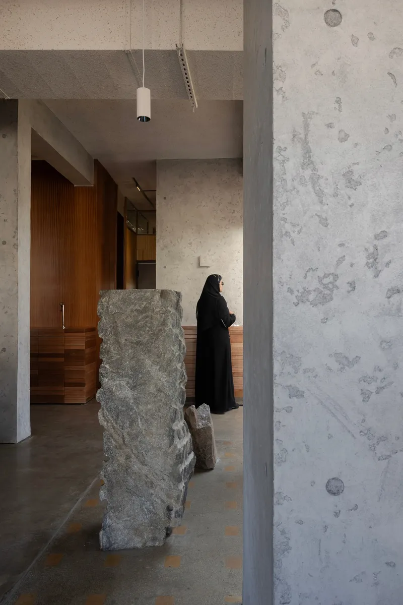Concrete column and textured wall beside a rough stone fragment with a person standing near timber millwork