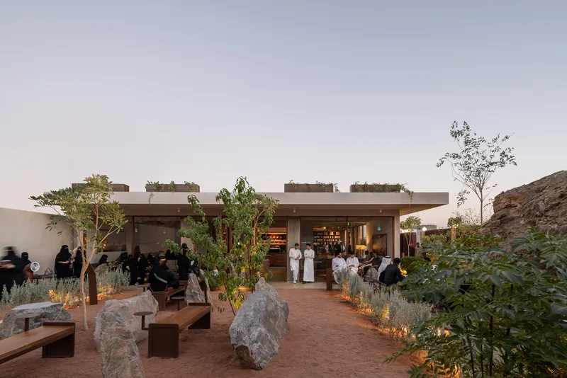 Outdoor terrace with planted roof openings, stone seating, and guests gathering at dusk beside a rocky hillside