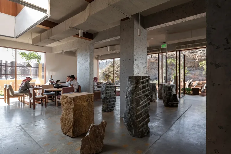 Open dining hall with carved stone column wraps, exposed concrete beams, and patrons seated near window walls framing the landscape
