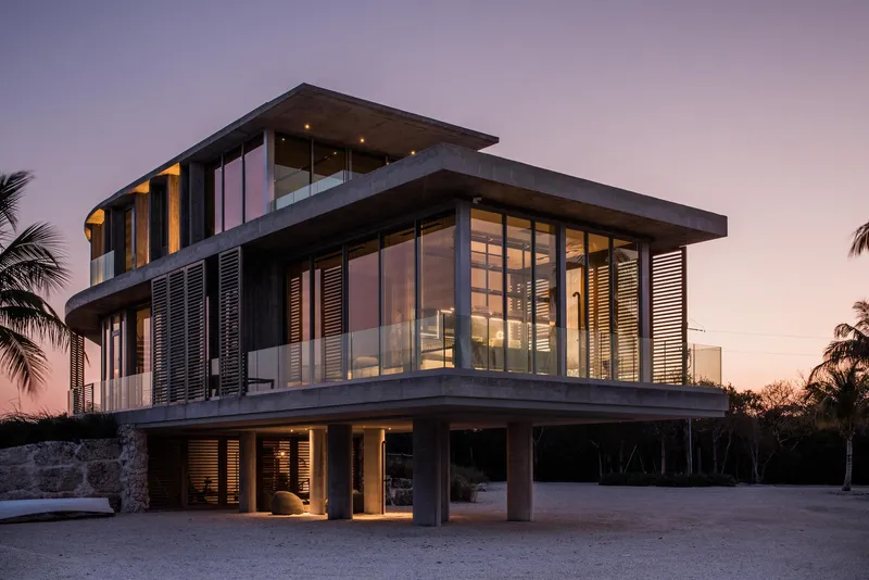Corner view of the three-level residence with illuminated interiors at dusk among palm trees